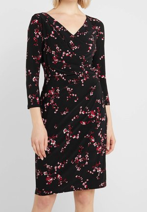 Kobieta nosząca czarną sukienkę do kolan z różowo-czerwonym wzorem kwiatowym i rękawami do łokcia, stojąca na jednolitym tle.