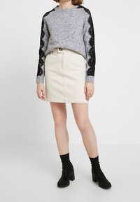 Maglione grigio a maglia con dettagli in pizzo nero sulle maniche, abbinato a una gonna in denim color crema e stivaletti neri con tacco.