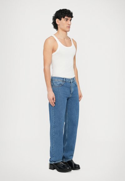 Bela majica brez rokavov, modre kavbojke z ravnimi hlačnicami in črne gležnjarice z visoko peto. Kavbojke imajo klasičen dizajn s petimi žepi in svetel pranje.