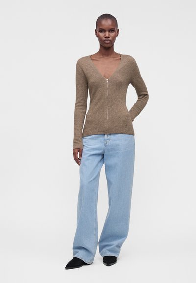 Femme portant un pull marron zippé, un jean bleu clair à jambes larges et des chaussures noires pointues, debout devant un fond clair uni.