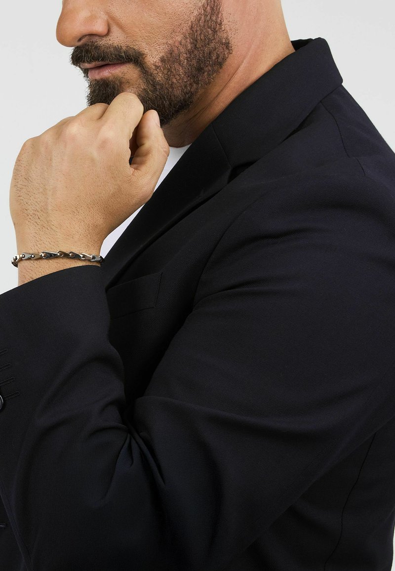 Giacca da abito nera su misura con una texture liscia, caratterizzata da spalle strutturate e una tasca sul petto. Un braccialetto metallico abbellisce il polso.