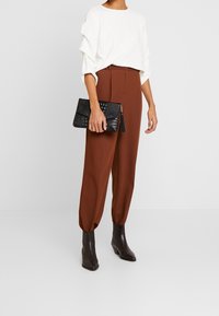 Kvinna i vit långärmad topp, högmidjade bruna byxor, mörka ankelstövlar, håller en svart texturerad clutchväska med fransen.