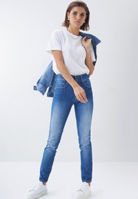 Jeans de mezclilla azul de cintura alta con un corte ajustado, que cuentan con bolsillos frontales y cierre de botón. Combinados con una camiseta blanca lisa y zapatillas.