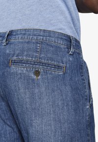 Primer plano de pantalones vaqueros azules con un bolsillo trasero abotonado, combinados con una camisa azul claro de textura ligera.