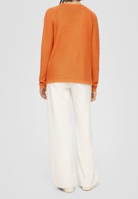 Oranger Strickpullover mit Rundhalsausschnitt und langen Ärmeln, kombiniert mit weißen, weit geschnittenen Hosen. Glatte Textur mit entspannter Passform.