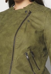 Kurtka z oliwkowej sztucznej zamszu z wysokim kołnierzem, ukośnym zamkiem błyskawicznym oraz dwoma ozdobnymi kieszeniami na zamek z srebrnymi okuciami.