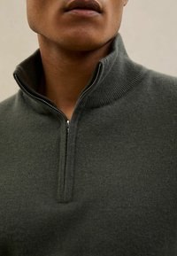 Tumši zaļš pus-pārklārta džemperis ar ribotu apkakli un rāvējslēdzēja detaļām. Audums izskatās mīksts un teksturēts, nodrošinot ērtu piegulumu.