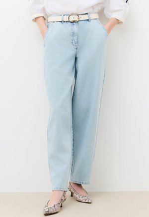 Jasnoniebieskie dżinsy o prostych nogawkach z białym paskiem, zestawione z butami na niskim obcasie w wzór skóry węża i ręce w kieszeniach.