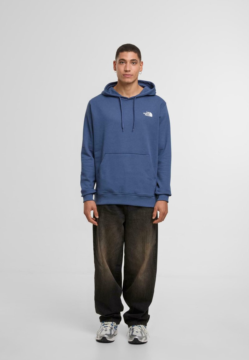 Marineblauwe hoodie met een voorzak en verstelbare trekkoorden, met een wit logo. Gecombineerd met wijde donkere jeans en sneakers.