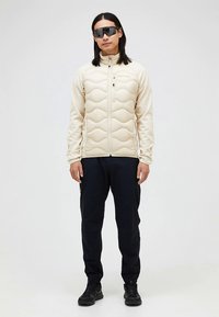 Beige dunjakke med hævede sømmønstre, lynlås foran; kombineret med mørke bukser og sorte sko. Tilbehør inkluderer store sorte solbriller.