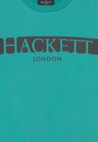 Maglietta in cotone turchese con un grande logo nero "HACKETT" e "LONDON" sotto, colletto rotondo e maniche corte.