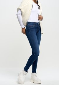 Jeans ajustados de mezclilla azul con una cintura alta, que cuentan con cinco bolsillos y detalles de costura sutiles, llevados con una blusa blanca de manga larga y un cárdigan de punto color crema.