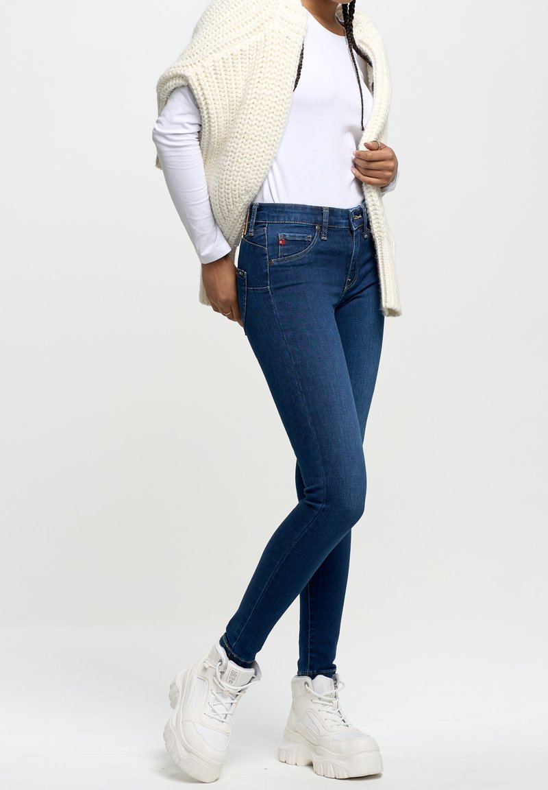 Jeans ajustados de mezclilla azul con una cintura alta, que cuentan con cinco bolsillos y detalles de costura sutiles, llevados con una blusa blanca de manga larga y un cárdigan de punto color crema.