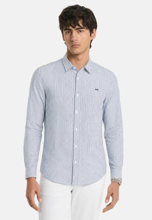 Giovane uomo che indossa una camicia a maniche lunghe con bottoni a righe verticali blu e bianche e pantaloni bianchi, in piedi davanti a uno sfondo semplice.
