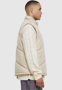 Beige pufferväst med hög krage, med quiltad textur och sidofickor, bärs över en gräddfärgad kabelstickad tröja, visad från sidan.
