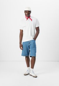 Weißes T-Shirt mit Druck, blaue Jeansshorts, rotes Halstuch, weiße Sneaker mit grauen Akzenten, hellfarbige Kappe. Minimalistisches Design.