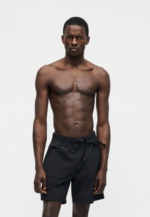 Μυώδης άνδρας σκουρόχρωμος στέκεται με το πρόσωπο προς τα εμπρός, φοράει μαύρο σορτς με ζώνη, μπροστά σε απλό φωτεινό φόντο.