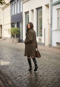Palton oversized, pufos, de culoare maro, cu guler, asortat cu bocanci din piele neagră, până la genunchi, și o geantă maro, pe o stradă cu pietre cubice.