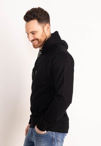 Felpa nera con cappuccio, tasca frontale, polsini a costine e vestibilità rilassata, abbinata a jeans blu chiaro. Texture liscia, senza motivi visibili.