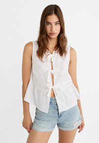 Biała bezrękawnikowa bluzka z trzema kokardkami z przodu, rozkloszowanym dołem i lekką, przewiewną fakturą. Połączona z jasnymi dżinsowymi szortami.
