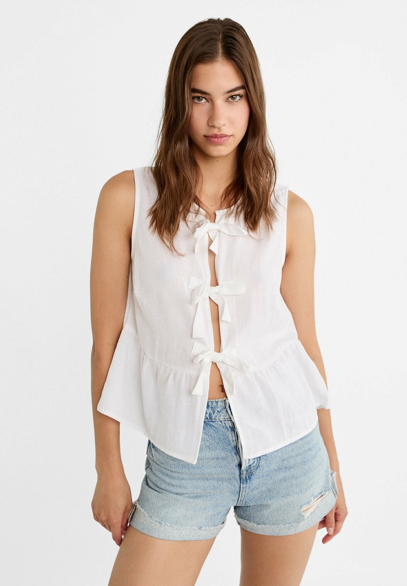 Biała bezrękawnikowa bluzka z trzema kokardkami z przodu, rozkloszowanym dołem i lekką, przewiewną fakturą. Połączona z jasnymi dżinsowymi szortami.