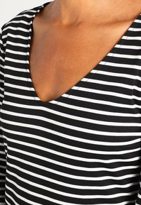 V-kauluksen muotoinen mustavalkoraitainen paita, joka on valmistettu pehmeästä puuvillakankaasta. Paita sisältää ohuita vuorotellen olevia raitoja ja siinä on sileä pinta.