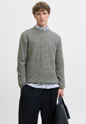 Grijze wollen trui met een ronde halslijn, ribbelmanchetten en -zoom, over een blauw-wit gestreept shirt heen. Zwarte tas in de hand.