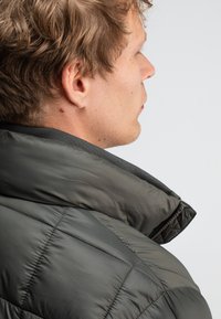Puffjacke aus dunkelgrauem Material mit hohem Kragen, gestepptem Textil und geschwungenen Nahtdetails an der Schulter.
