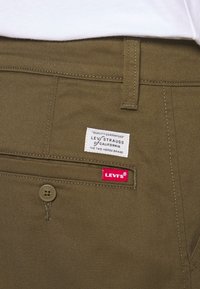 Olivově zelené bavlněné kalhoty s brandovanou nášivkou "Levi Strauss" a červenou štítkem "Levi's". Obsahují zapínání na knoflík a prošité švy.