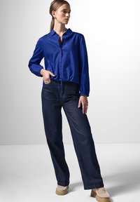Camicia button-up a maniche lunghe di un blu reale, con orlo a cordoncino, abbinata a jeans a gamba larga blu scuro e scarpe beige senza lacci.