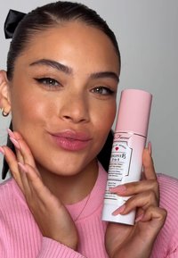 Witte sprayfles met roze dop, gelabeld "Hangover 3-in-1". Model met donker haar en lichtroze nagels poseert in een roze trui.