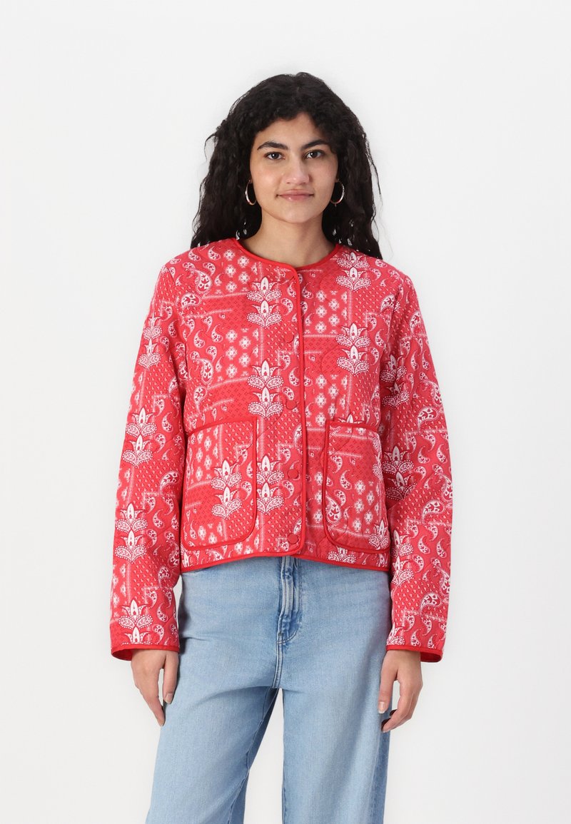 Rote gesteppte Jacke mit Blumen- und Paisleymuster, runder Halsauschnitt, Knopfverschluss und Fronttaschen. Kombiniert mit hellblauen Jeans.