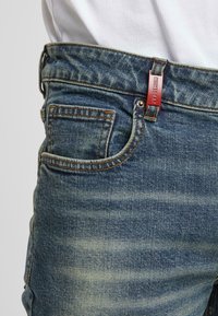 Džínové kalhoty ve středně modré barvě s vybledlým vzhledem, se jednou přední kapsou, měděným šitím a červenou značkovou nášivkou na pasu.