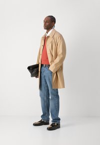 Beige lichtgewicht trenchcoat over een oranje trui, blauwe jeans en zwarte schoenen met versieringen. Houdt een zwarte tas vast.