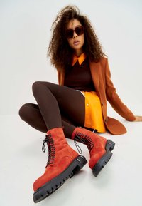 Rode suede veterschoenen met een stevige zwarte zool en decoratieve vetersluiting. Het model draagt een bruine top, een oranje shirt en een bruine panty.