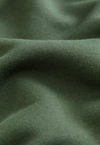 Gros plan sur un tissu vert doux et texturé avec un fin motif tricoté et des plis légers créant des effets de lumière et d'ombre.