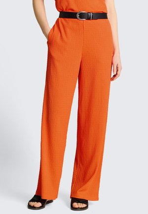 Donna che indossa pantaloni larghi arancioni vivaci e strutturati con cintura nera e sandali neri aperti in punta, una mano in tasca, in piedi davanti a uno sfondo semplice.