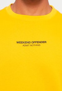Yellow cotton t-shirt featuring a round neckline, short sleeves, and printed text: "WEEKEND OFFENDER" and "ADMIT NOTHING" in black.