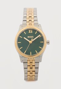 Ανδρικό ρολόι χειρός με πράσινο καντράν, χρυσούς δείκτες ώρας, βραχιόλι από ασημί και χρυσά μεταλλικά κρίκους, και λογότυπο "BOSS" κάτω από τις 12 η ώρα.