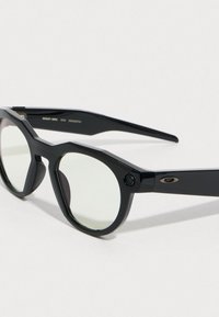 Mustat aurinkolasit kulmikkaalla muotoilulla, joissa on pyöreät linssit ja näkyvä logo sivussa. Teksturoitu muovikehys ja hienovaraiset yksityiskohdat.