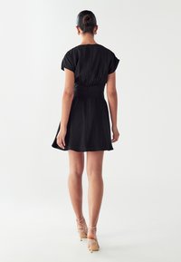 Woman with hair in a bun wearing gold hoop earrings, a short black dress, and beige high-heeled sandals, standing with back facing forward.