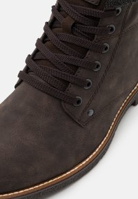 Stivale in pelle marrone con superficie testurizzata, lacci marrone scuro e occhielli in metallo nero. Cuciture bianche lungo la suola e suola in gomma nera.