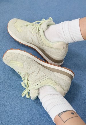 Pieds portant des chaussettes blanches et des baskets New Balance de couleur claire sur une surface bleue texturée, montrant un petit tatouage rectangulaire sur une cheville.