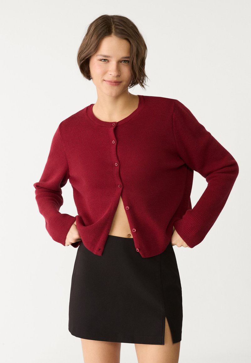 Roter gerippter Cardigan mit rundem Ausschnitt und vorderer Knopfleiste, kombiniert mit einem schwarzen Miniröckchen mit seitlichem Schlitz.