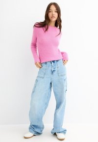 Una joven con cabello largo que lleva un suéter rosa de felpa, unos pantalones cargo sueltos de color azul claro y zapatillas blancas, de pie con las manos en los bolsillos.