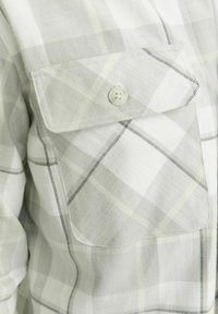 Camisa de franela gris claro a cuadros que presenta un bolsillo en el pecho con solapa y botón, dobladillo curvado y líneas oscuras contrastantes en el patrón.