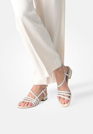 Feet wearing beige open-toe strappy sandals with low block heels and white wide-leg pants against a plain background.
