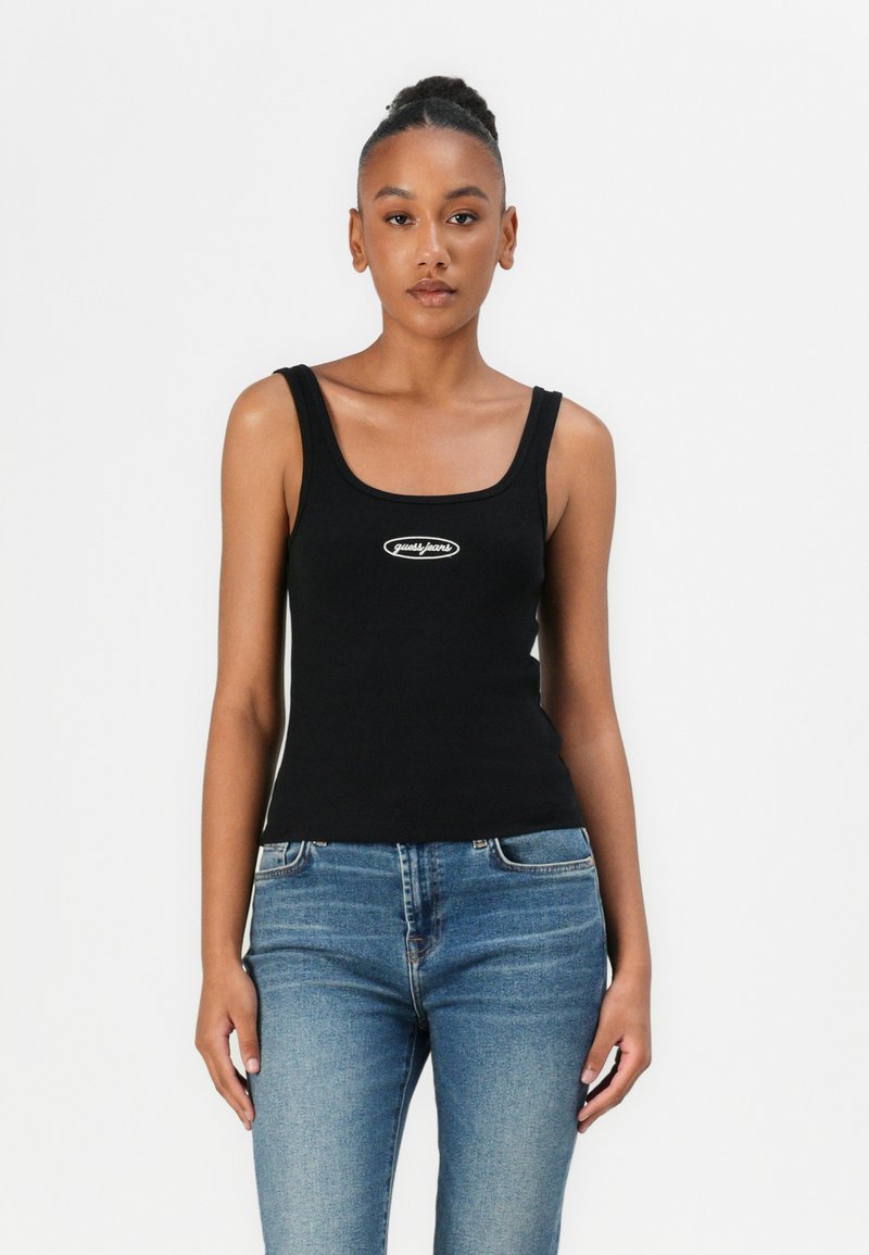 Schwarzes geripptes Tanktop mit quadratischem Ausschnitt und einem kleinen weißen Logo auf der Brust, kombiniert mit hellblauen Jeans.