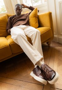 Braune Lederschuhe mit einer strukturierten Oberseite und elastischem Detail, getragen zu cremefarbenen Hosen, sitzend auf einem gelben Sofa mit Holzboden.