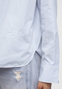 Chemise rayée bleu clair et blanche avec "Hugo" brodé près de la taille, portée avec un jean déchiré bleu clair.